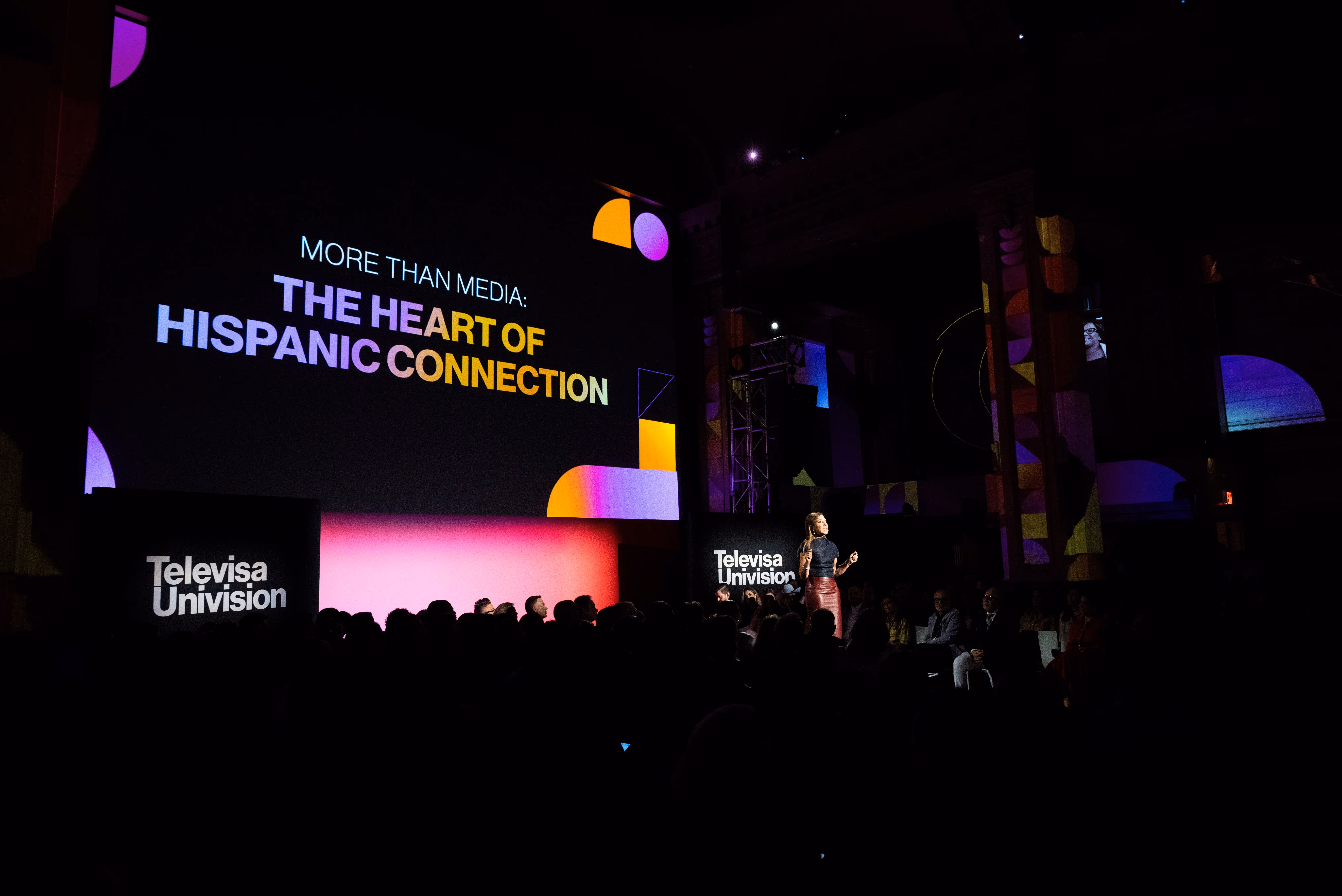 Woman speaking on stage at an event with a large screen behind displaying the text 'More than media: The heart of Hispanic connection' and Televisa Univision logos.