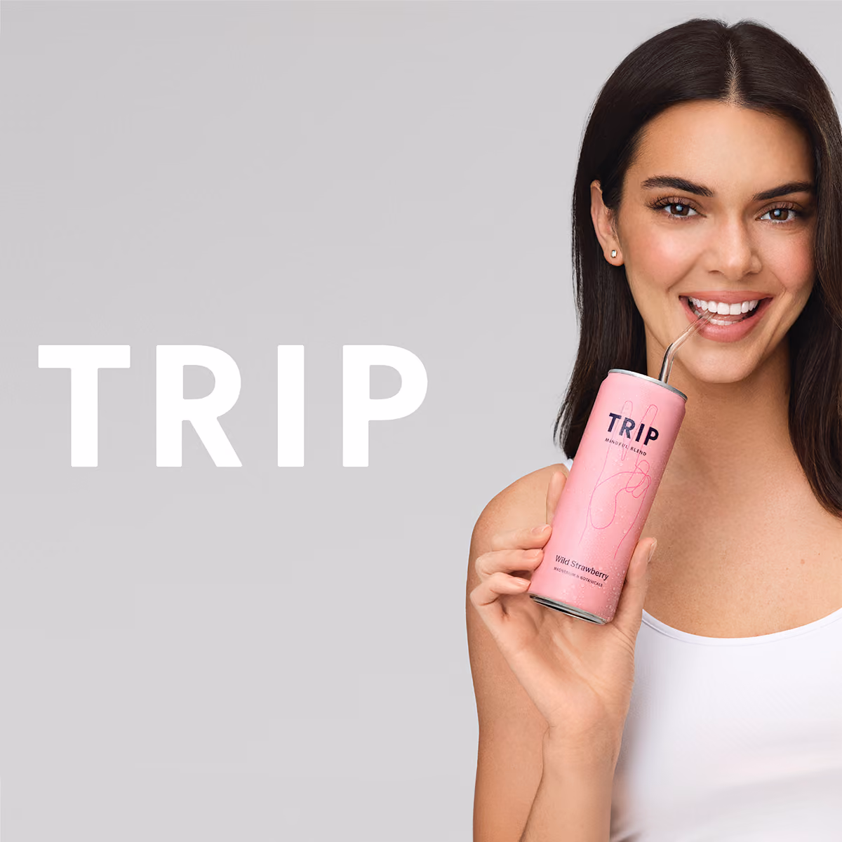 Kylie Jenner with long dark hair holds a pink TRIP Wild Strawberry drink can and sips through a metal straw against a light gray background, with large white “TRIP” text on the left.