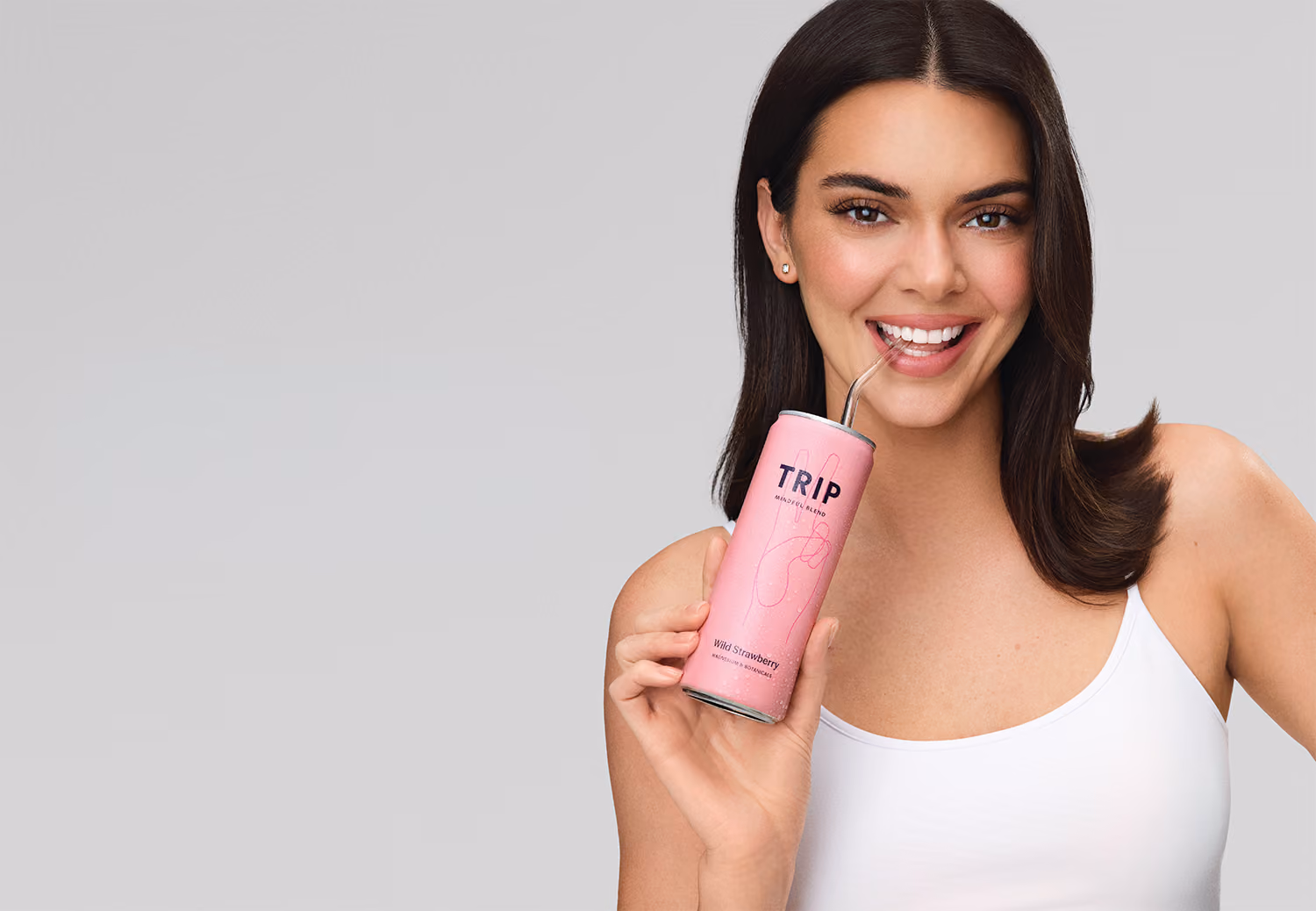 Kylie Jenner with long dark hair holds a pink TRIP Wild Strawberry drink can and sips through a metal straw against a light gray background, with large white “TRIP” text on the left.
