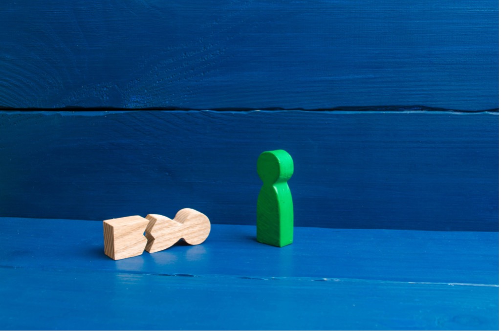 A green wooden human figure stands beside a natural wood human figure broken into two pieces on a blue wooden surface.