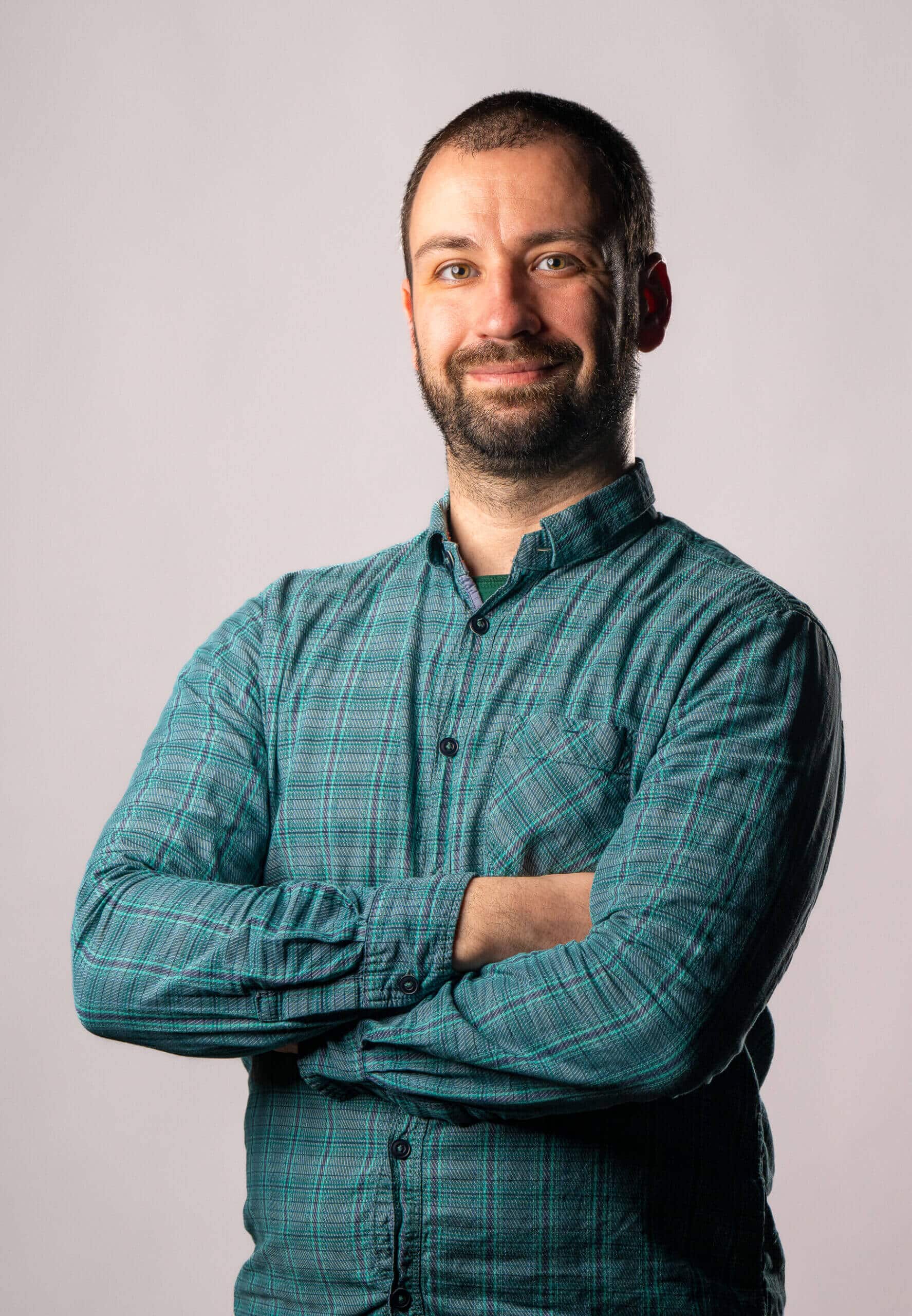 Smiling man with short hair and beard wearing a green plaid shirt with arms crossed against a plain background.
