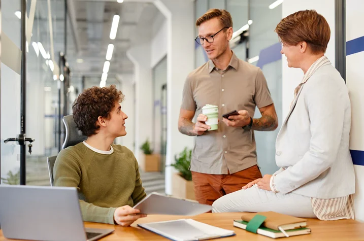 Three colleagues talking in office, reviewing documents and discussing tasks during employee onboarding or workplace training session