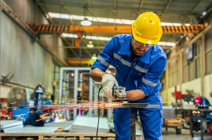 Worker in protective gear using angle grinder in factory workshop, demonstrating safe tool handling during industrial training or compliance work