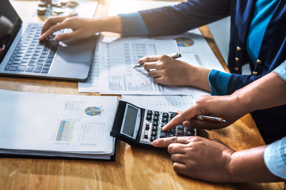 Two people working with a laptop, calculator, and financial documents with charts on a wooden table.
