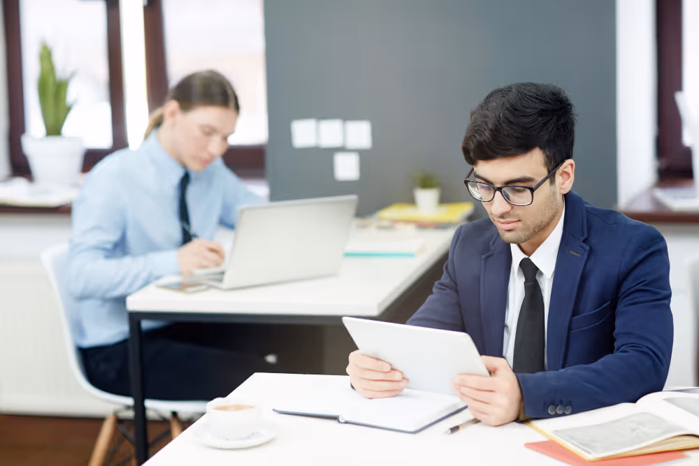 Two professionals working in an office, one reading a tablet and the other writing near a laptop.