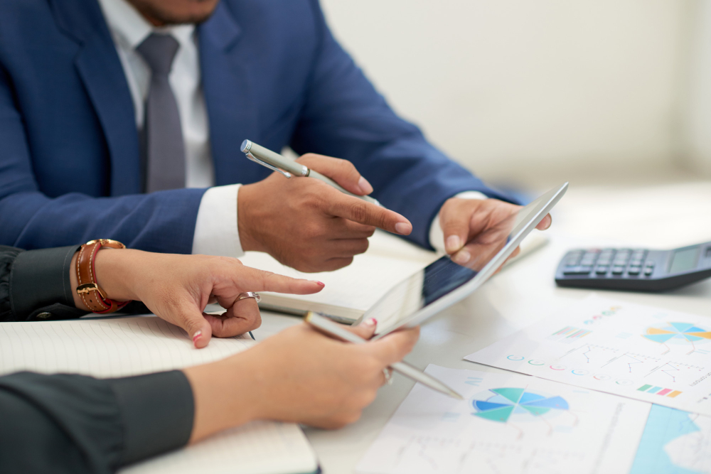 Two business professionals pointing at a tablet screen during a discussion, with financial charts and a calculator on the table.
