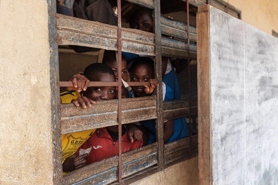 Mehrere afrikanische Kinder schauen lachend und neugierig durch ein vergittertes Metallfenster in einem Klassenzimmer; im Vordergrund steht eine alte Tafel.