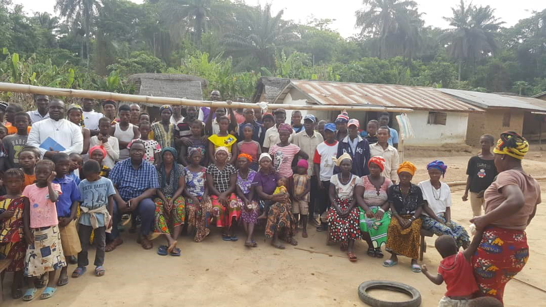 Große Gruppe von Frauen, Männern und Kindern steht und sitzt für ein Gruppenfoto in einem ländlichen afrikanischen Dorf; im Hintergrund einfache Häuser mit Wellblechdächern und Palmen.
