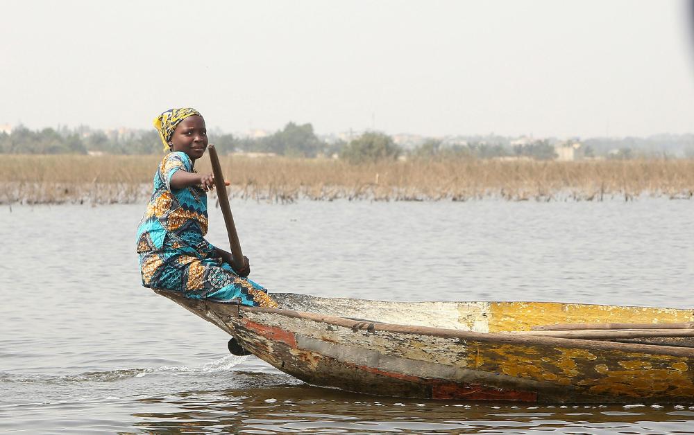 Ein Mädchen in traditioneller Kleidung paddelt ein Holzboot über einen Fluss in Benin