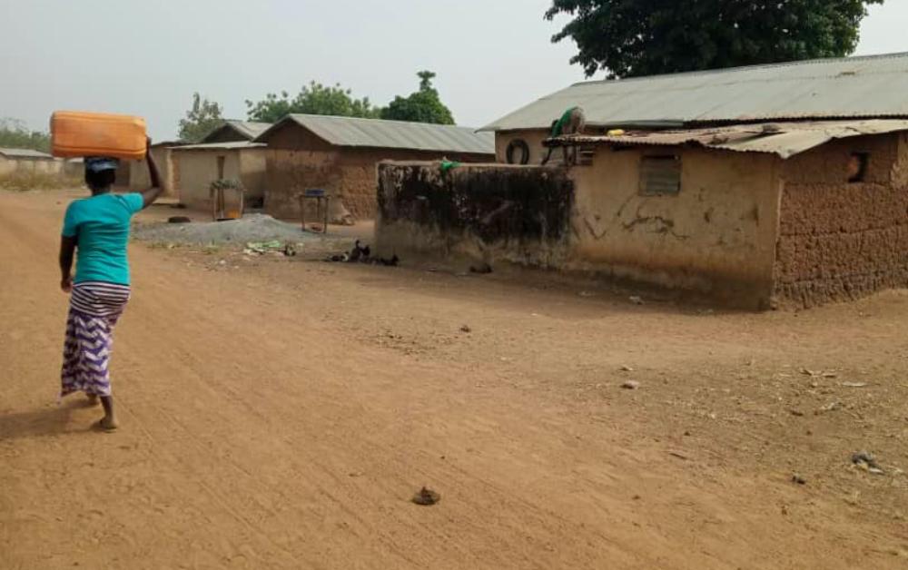 Eine Frau trägt ein schweres Paket auf dem Kopf und geht durch ein schmutziges Dorf in Benin