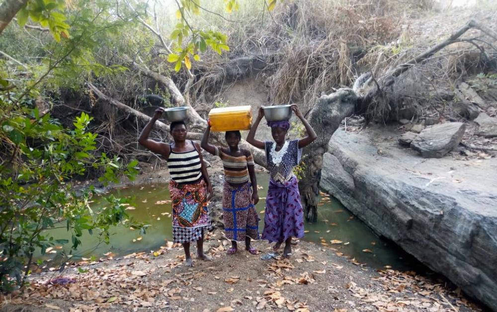 3 Frauen in Benin vor einer schmutzigen Wasserstelle mit Wasserkanistern auf den Köpfen