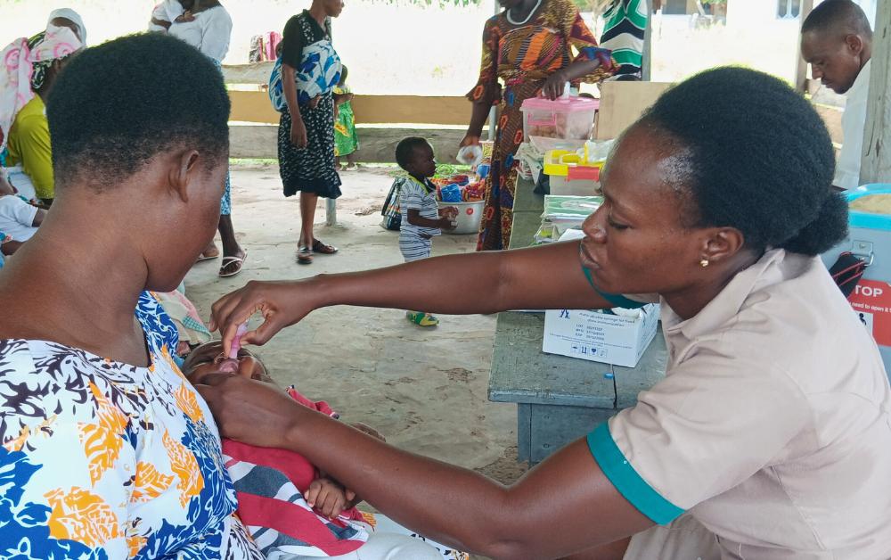 Afrikanische Krankenschwester verabreicht einem Kleinkind eine orale Impfung, während das Kind auf dem Schoß der Mutter sitzt; im Hintergrund weitere Frauen und Kinder in einer offenen Gesundheitsstation.