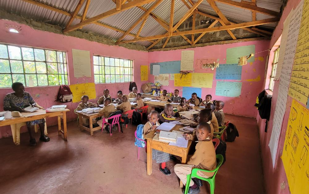 Afrikanisches Klassenzimmer mit pink gestrichenen Wänden; zahlreiche Grundschulkinder sitzen an kleinen Tischen mit Schulheften, eine Lehrerin sitzt vorne links. Die Wände sind mit Lernplakaten und Schreibübungen bedeckt.
