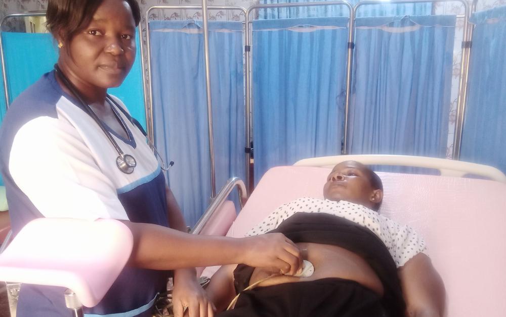 Afrikanische Krankenschwester untersucht mit einem Stethoskop den Bauch einer schwangeren Frau, die in einem Klinikbett liegt; im Hintergrund Sichtschutzwände in einem einfachen Untersuchungsraum.
