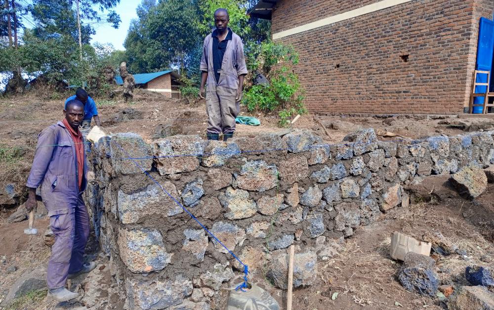 Drei Männer arbeiten an einem Rohbau aus Natursteinen in einer hügeligen Gegend in Afrika; einer der Männer hält Werkzeug in der Hand, im Hintergrund steht ein Gebäude mit Ziegelfassade und blau gestrichener Tür.