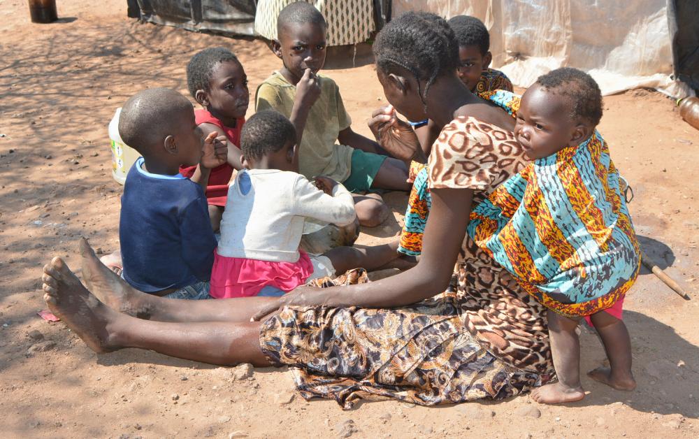 Afrikanische Frau sitzt barfuß auf dem Boden und umringt von mehreren kleinen Kindern, eines trägt sie auf dem Rücken; die Gruppe befindet sich auf trockenem, staubigem Untergrund vor einfachen Unterkünften.