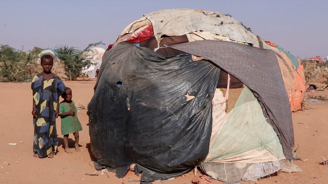 Zwei afrikanische Kinder stehen barfuß im Sand vor einer notdürftig errichteten Hütte aus Plastikplanen, Stofffetzen und Decken in einem Flüchtlingslager in trockener Wüstenlandschaft.