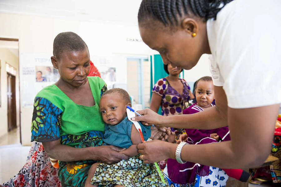 Afrikanische Mutter hält ihr Kleinkind auf dem Schoß, während eine Mitarbeiterin in einem Gesundheitszentrum den Oberarmumfang des Kindes mit einem Messband zur Feststellung von Mangelernährung misst; im Hintergrund weitere Frauen mit Kindern.