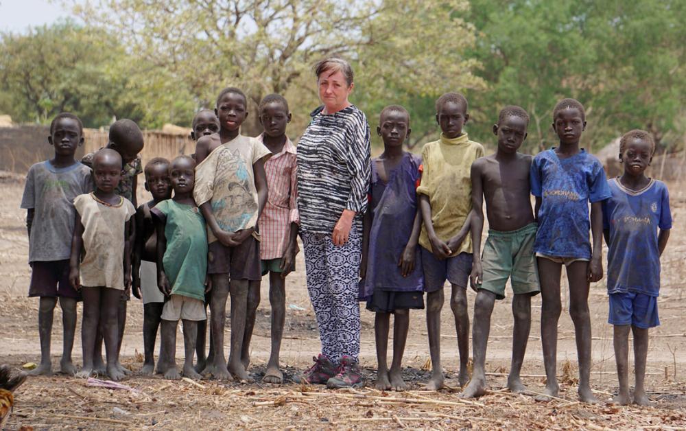Gruppe afrikanischer Kinder in zerschlissener Kleidung steht barfuß im Trockengebiet des Südsudan neben einer europäischen Helferin; im Hintergrund Bäume und einfache Lehmhütten.