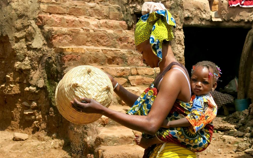 Mutter mit Baby auf dem Rücken entleert einen Korb – Nahrungssicherheit und Mutterrolle in Burkina Faso.