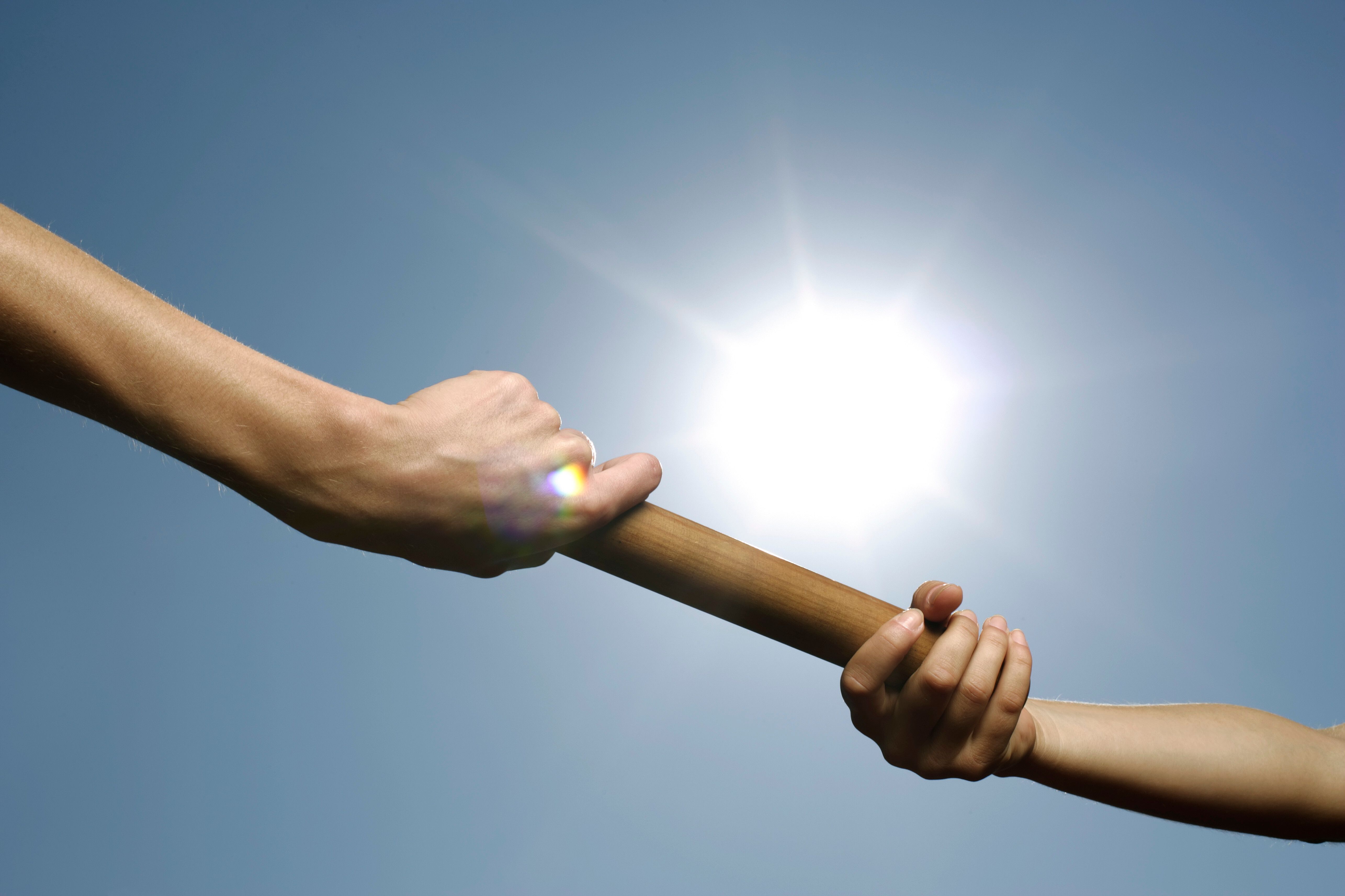 Two hands passing a baton in a relay race against a bright sky, symbolizing a business handoff or succession
