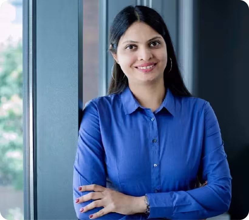 Smiling woman with long black hair wearing a blue button-up shirt, standing by a window with arms crossed.