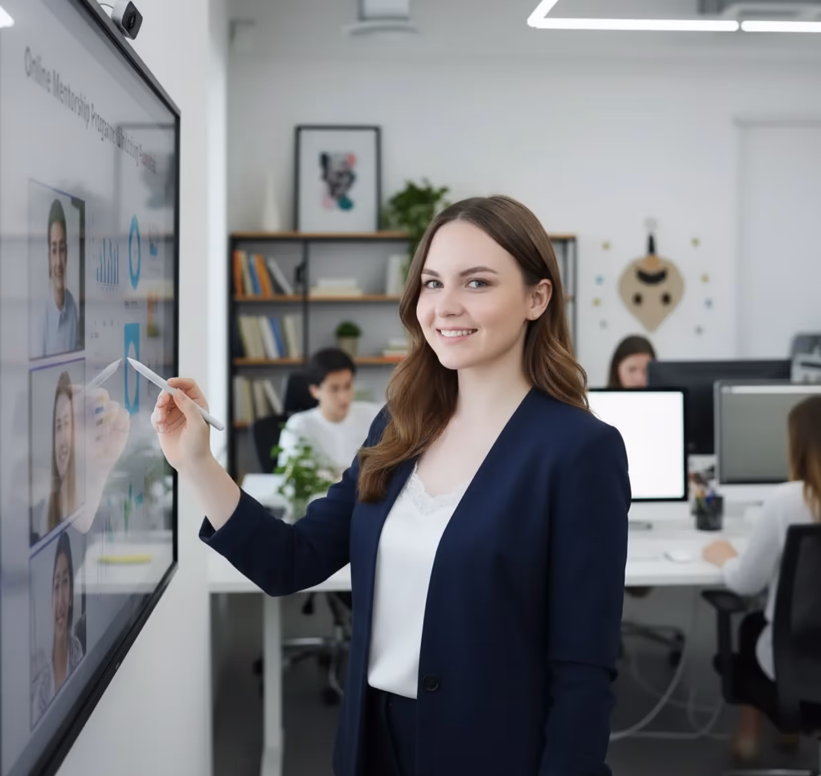 Smiling businesswoman using a stylus to point at a large touch screen in a modern office with colleagues working in the background.