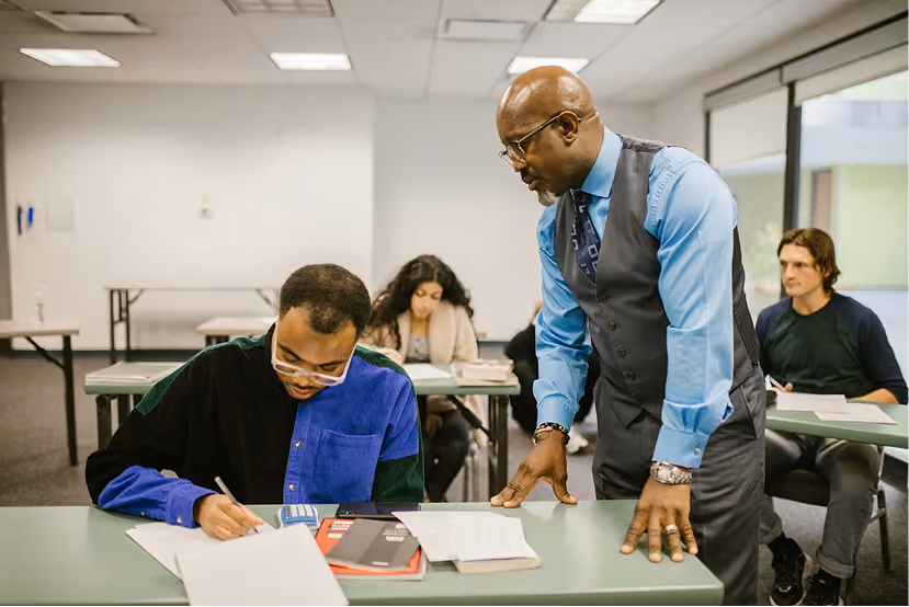 Teacher in vest and tie helps student writing at desk in classroom with other students seated behind.
