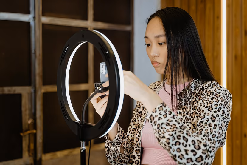 Woman adjusting smartphone on ring light for recording or streaming in an indoor setting.