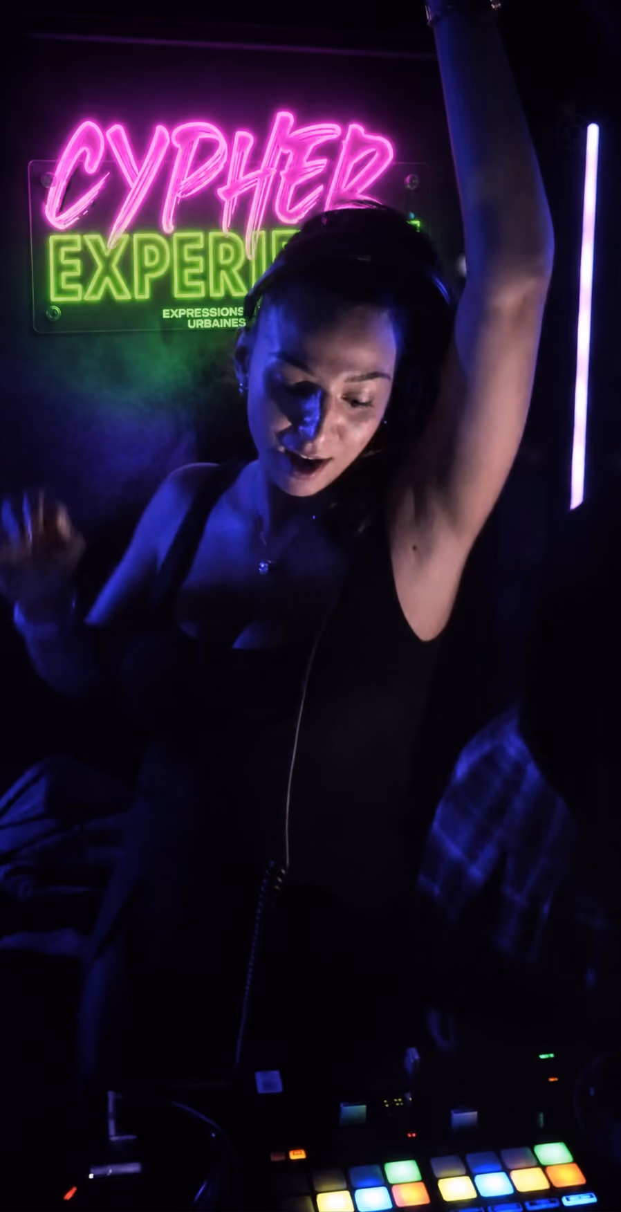 Une DJ femme s'amuse dans une ambiance nocturne avec un panneau néon «CYPHER EXPERIENCE» en arrière-plan.