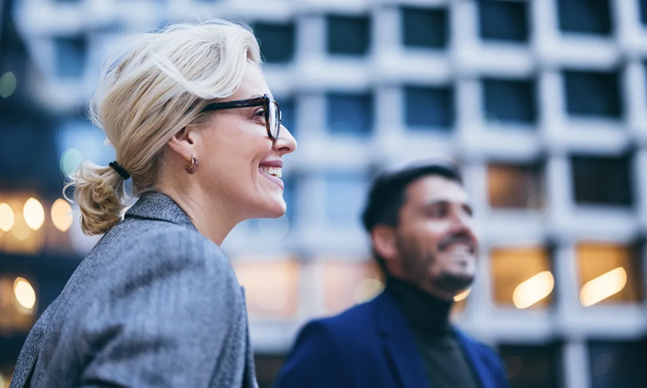 Femme blonde souriante portant des lunettes et un blazer gris, avec un homme flou en arrière-plan devant un bâtiment moderne.