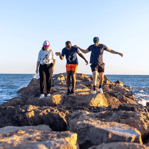 Trois personnes marchent en équilibre sur des rochers au bord de la mer au coucher du soleil.