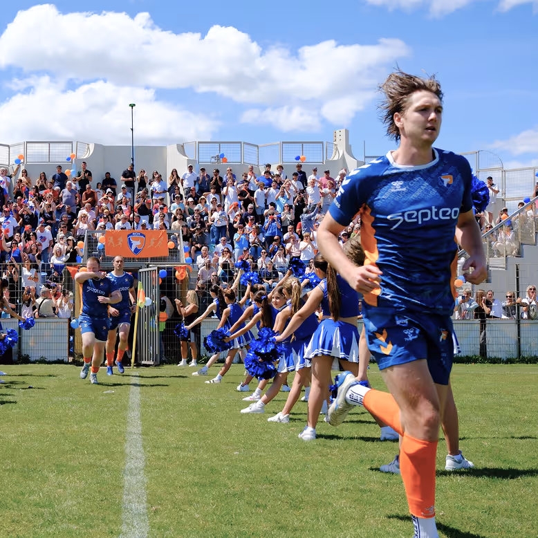 Joueurs de rugby courant sur le terrain devant des pom-pom girls et une foule enthousiaste dans les gradins.