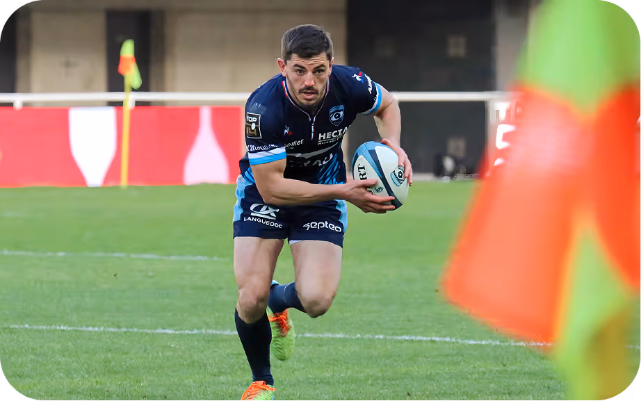 Joueur de rugby en maillot bleu foncé tenant un ballon ovale lors d'un match sur le terrain.