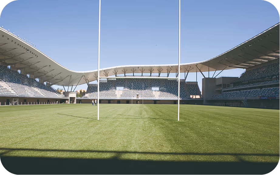 Terrain de rugby vide avec poteaux de but et tribunes couvertes sous ciel bleu clair.