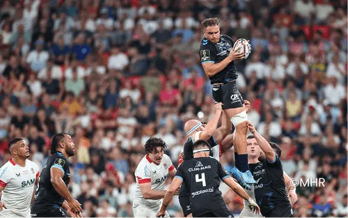 Joueur de rugby en maillot bleu foncé tenant un ballon ovale lors d'un match sur le terrain.