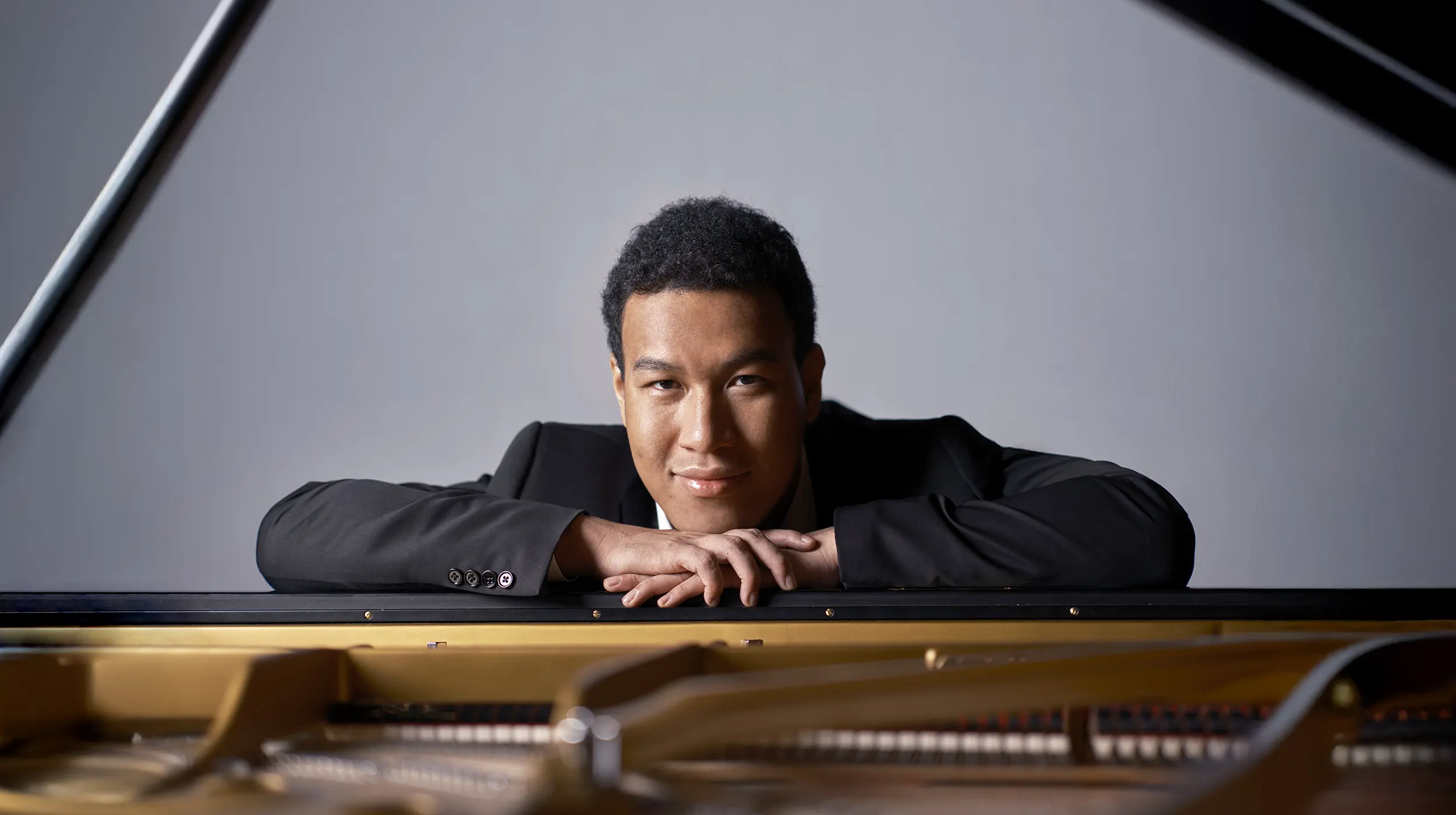 Photo of Clayton sitting in front of the piano, arms crossed leaning on top of the piano frame