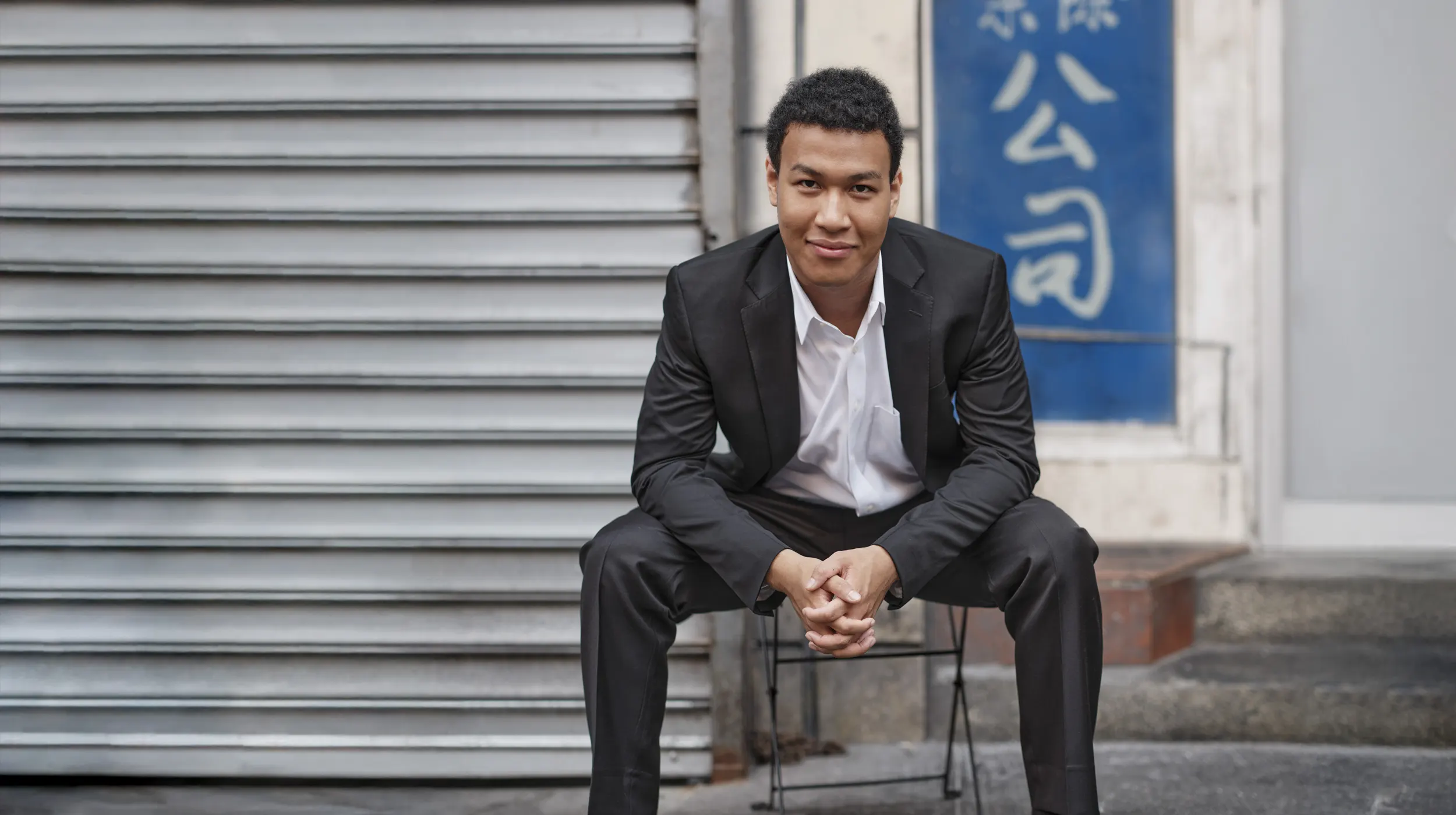 Photo of Clayton leaning forward in a chair on the street in front of a metal door