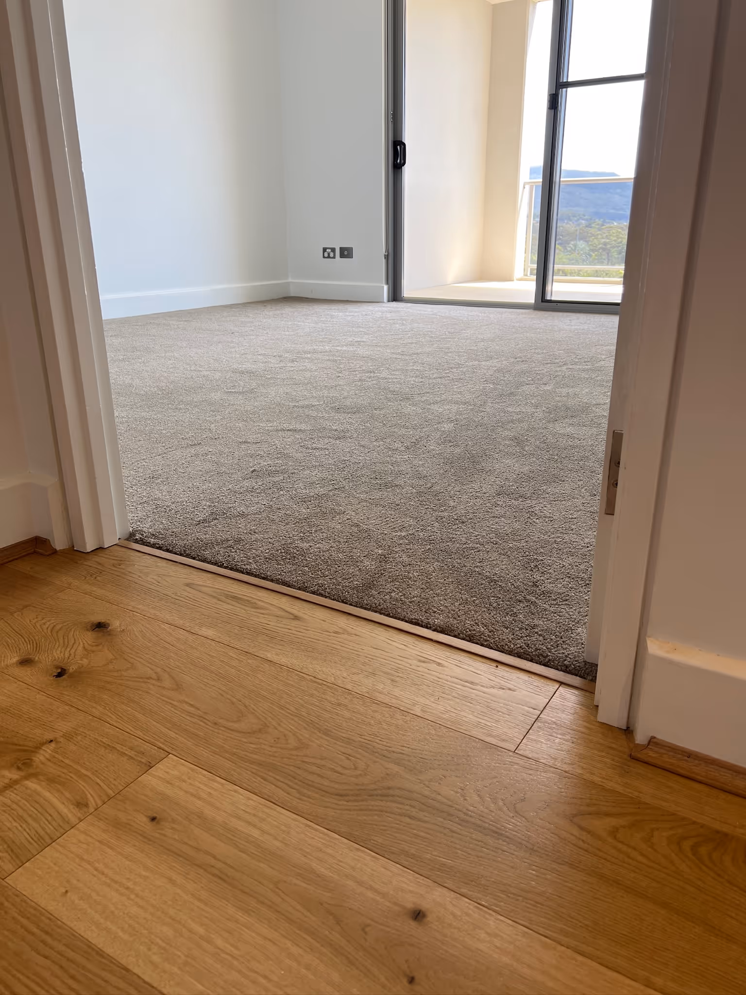 Hallway with timber flooring leading into a bedroom with carpet flooring. The transition highlights the contrast between the warm wood tones and the soft, textured carpet.