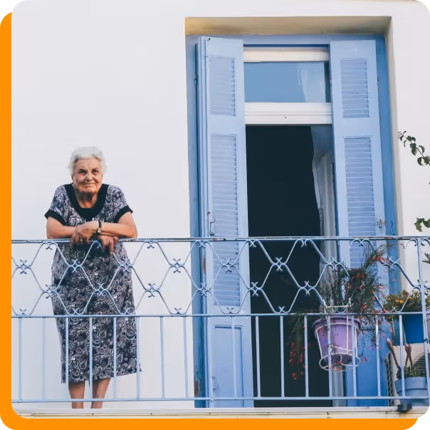 Elderly woman standing on a balcony with blue shutters and potted plants.