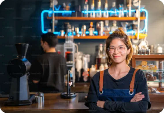 Smiling young female barista with glasses and apron standing with arms crossed in a coffee shop with shelves of bottles and a coffee grinder behind her.