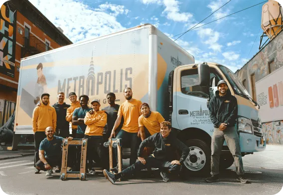 Group of nine delivery workers posing in front of a Metropolis Moving company truck on a sunny day.