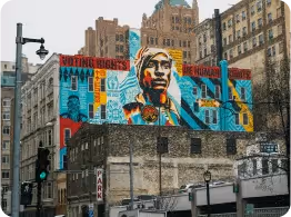 Colorful mural of a Native American man in traditional headdress painted on a building in an urban cityscape.