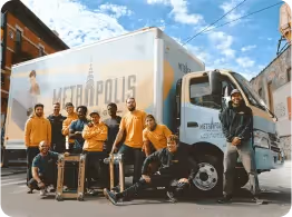 Group of twelve people posing casually in front of a white delivery truck with the word 'Metropolis' on its side, outdoors on a sunny day.