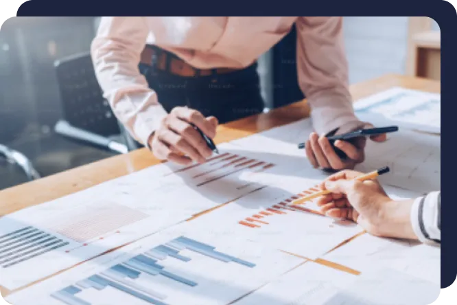 Two people reviewing and discussing printed charts and graphs on a desk, one holding a pen and the other a phone.