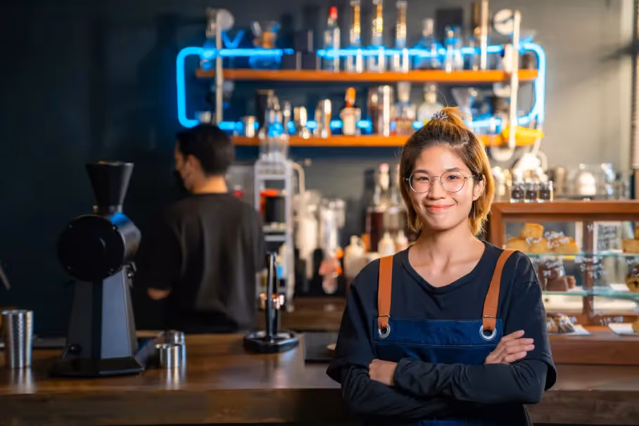 Young woman wearing glasses and an apron standing confidently with arms crossed in a coffee shop with a barista working behind the counter.