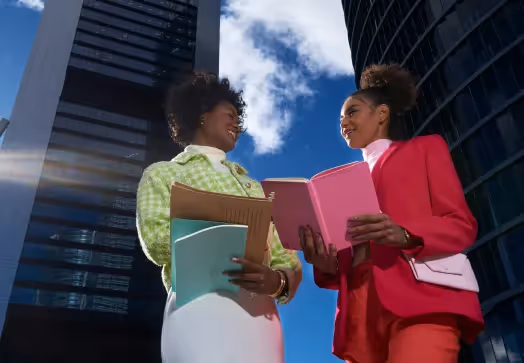 Two professional women holding folders and smiling while standing outdoors with modern skyscrapers and blue sky in the background.