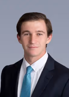 Young man with short brown hair wearing a dark suit, white shirt, and light blue tie against a plain gray background.