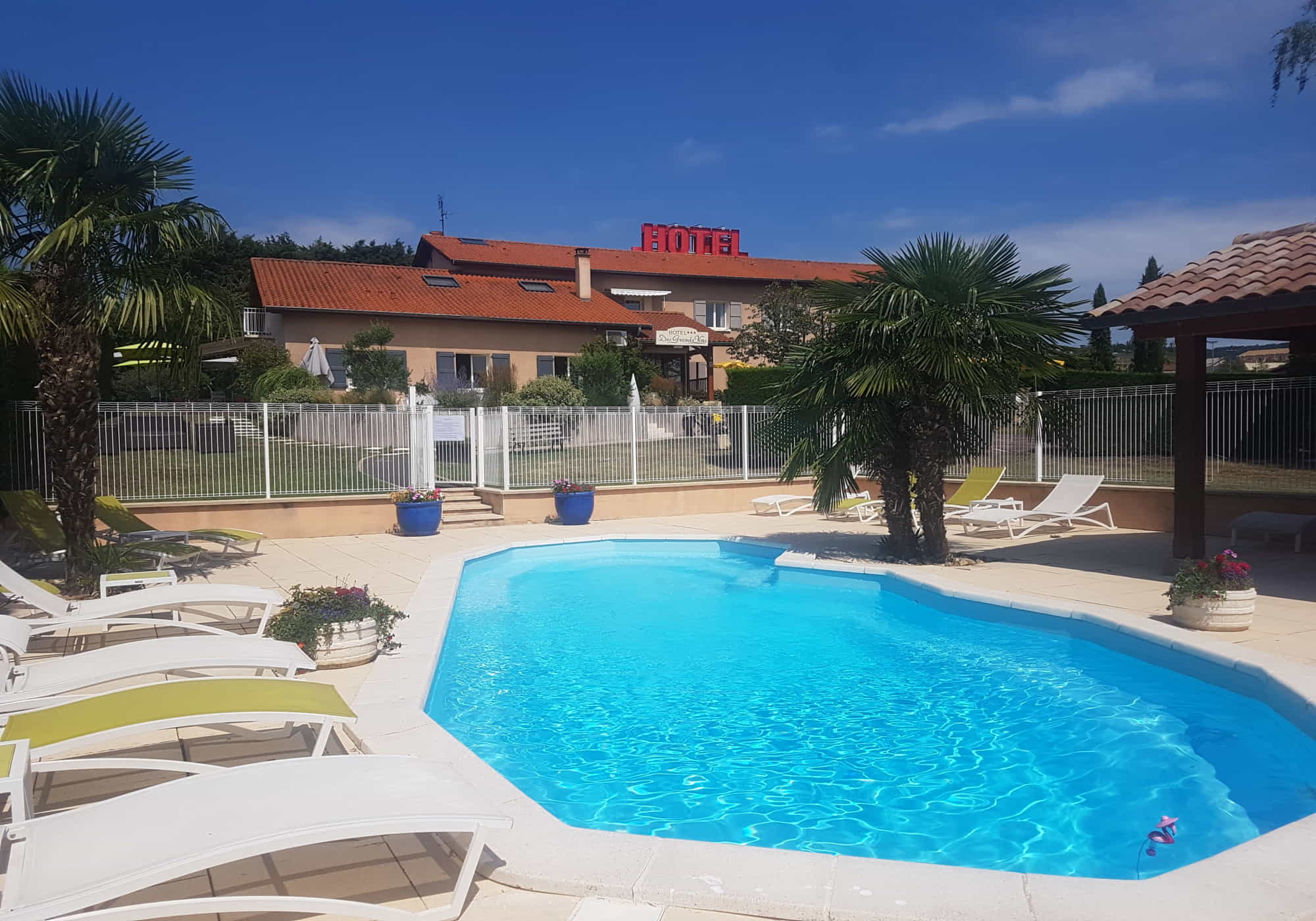 Piscine extérieure avec transats et palmiers devant un hôtel sous ciel bleu.