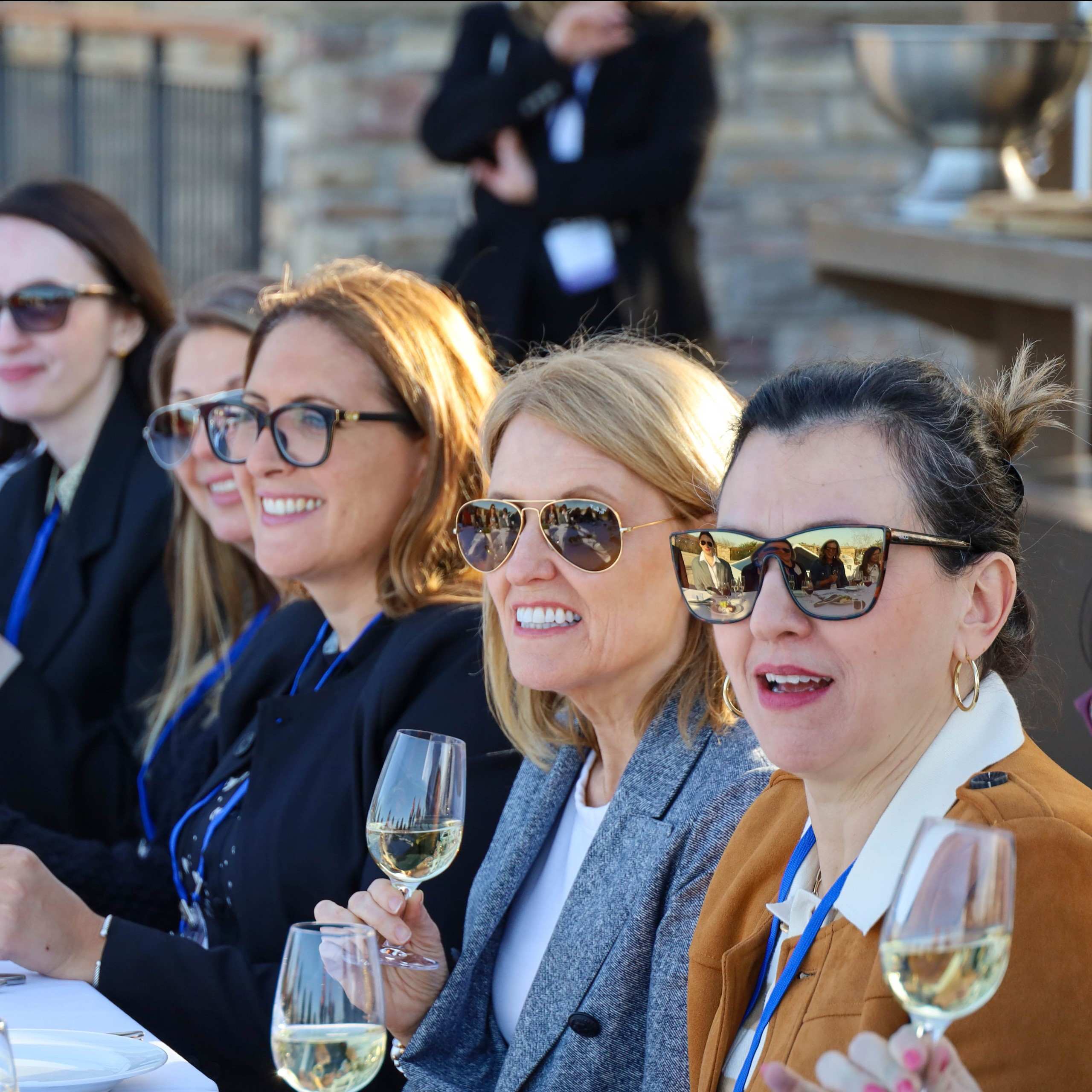Attendees wearing sunglasses share smiles around an outdoor dinner table